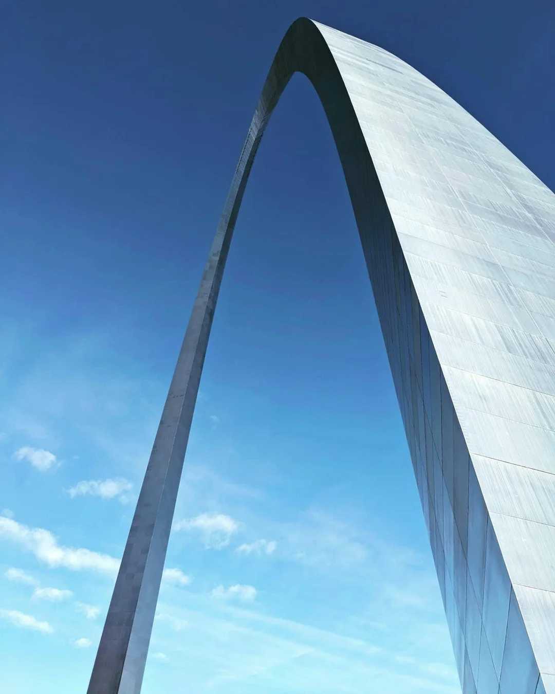 The image shows the Gateway Arch in St. Louis, Missouri, rising against a bright blue sky with wispy clouds. The photo captures the arch from a low angle, emphasizing its gleaming metallic surface and sweeping curve.