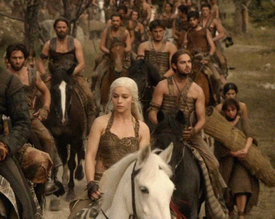 A woman with long, white-blonde hair rides a white horse at the front of a group of people on horseback, all dressed in brown, rustic clothing in a forest setting.