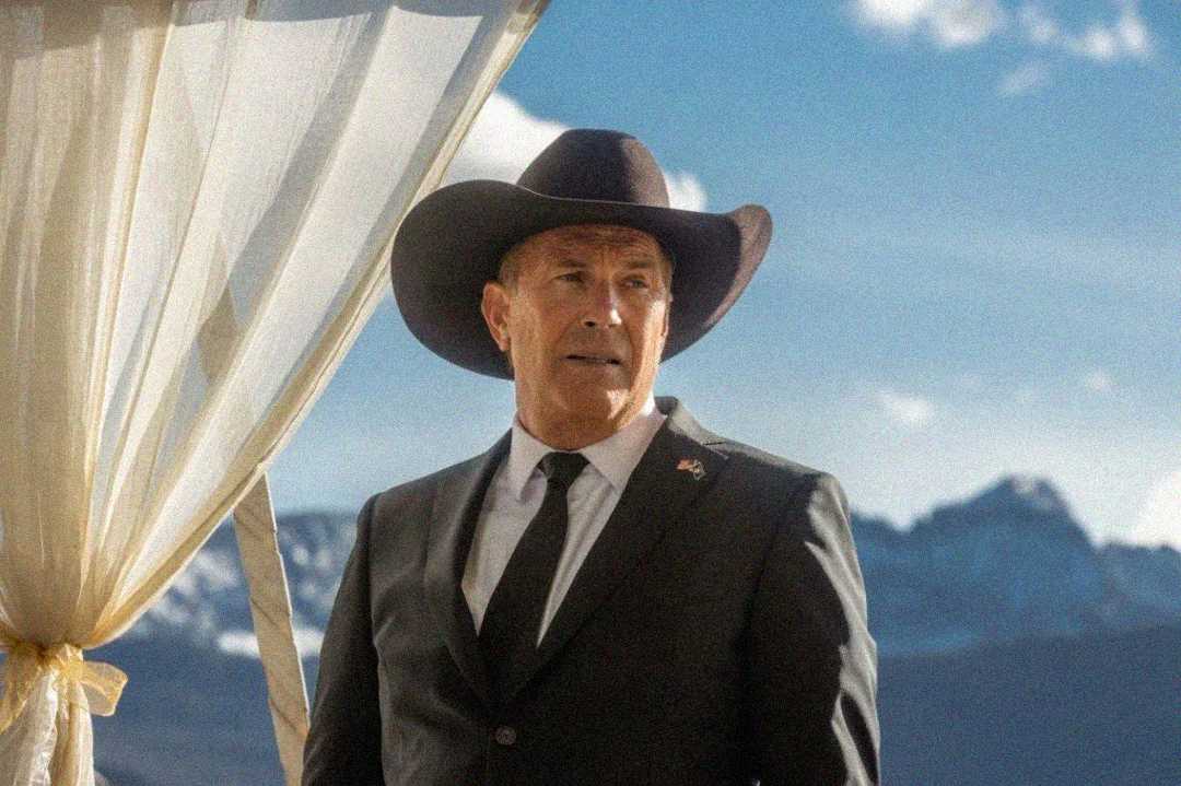 A man in a black cowboy hat and suit stands outdoors beside a white curtain, with mountains and a blue sky in the background.