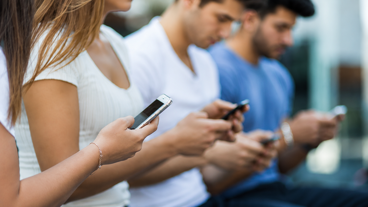 Four young adults sit in a row outdoors, each focused on their smartphones. The image is slightly blurred, emphasizing their concentration on their devices rather than their surroundings.