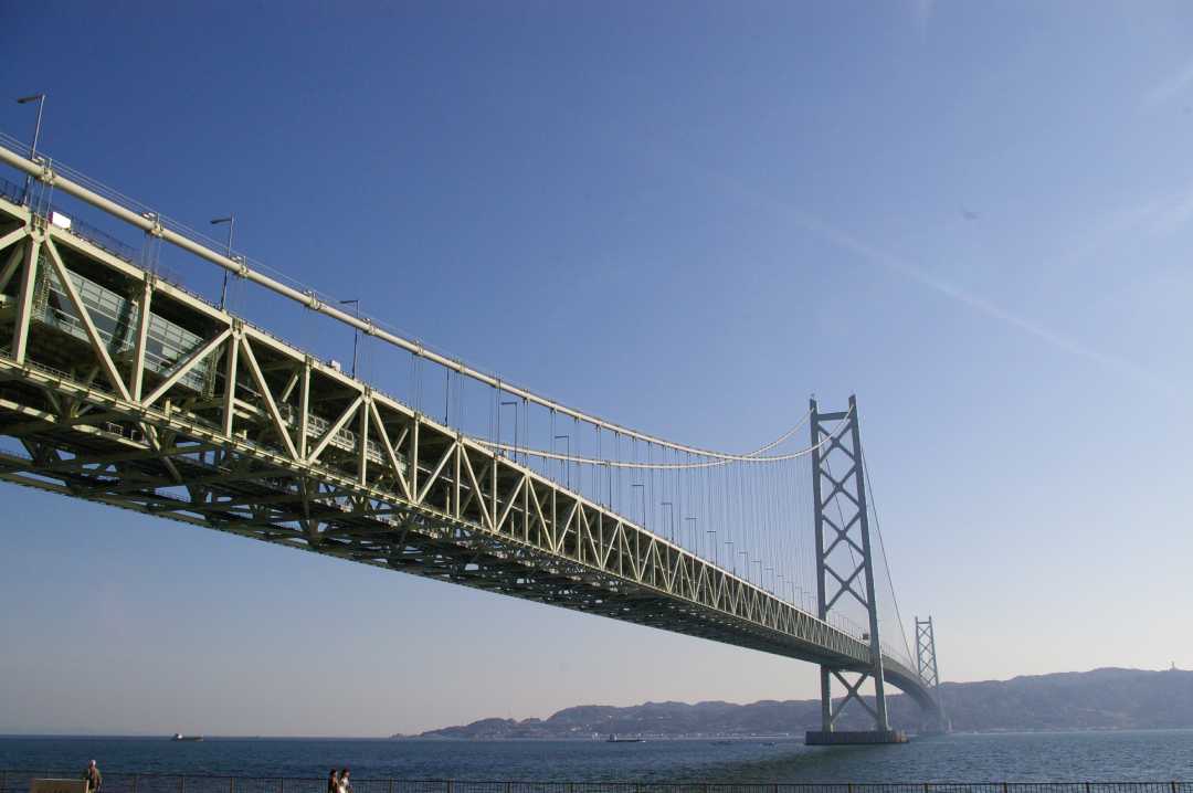 A large suspension bridge spans across a body of water, with clear blue sky above and distant hills visible in the background. A few people are standing near the waterfront below the bridge.