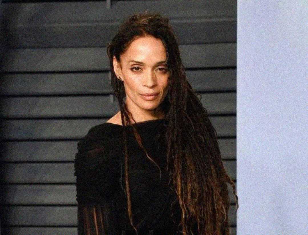 A woman with long, dark dreadlocks wearing a black top stands in front of a dark, striped background, looking at the camera with a neutral expression.