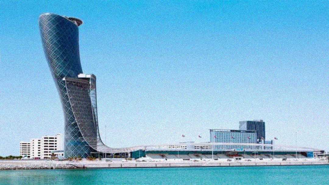 A modern, uniquely curved skyscraper stands beside a waterfront under a clear blue sky, with smaller buildings and palm trees nearby. The tower leans dramatically to one side.