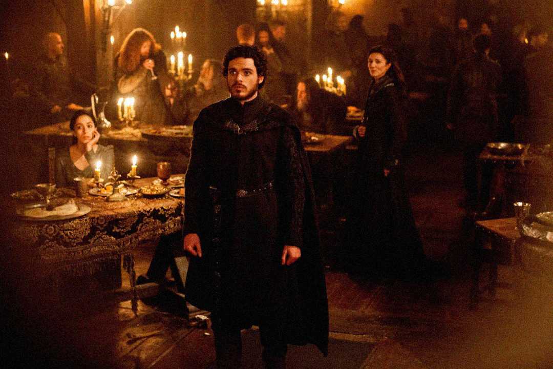 A man in dark medieval clothing stands in a candle-lit hall, surrounded by people sitting and standing near tables filled with food. The atmosphere is tense and dramatic, with dim, warm lighting.