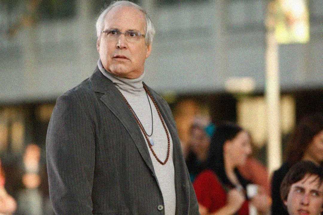 An older man with gray hair and glasses, wearing a gray suit jacket, light gray turtleneck, and a beaded necklace, stands outdoors looking to the side with a concerned expression. Blurred people are in the background.