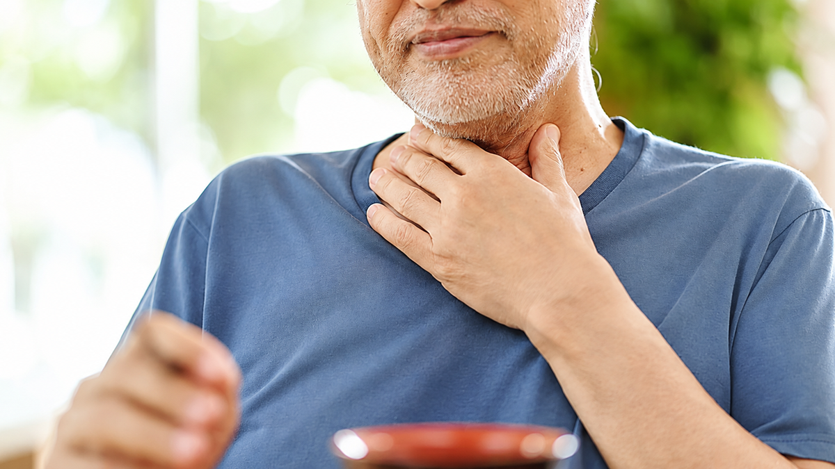 An older man wearing a blue shirt touches his throat with a pained expression, suggesting discomfort or a sore throat. He is sitting indoors with a bowl in front of him and greenery in the background.