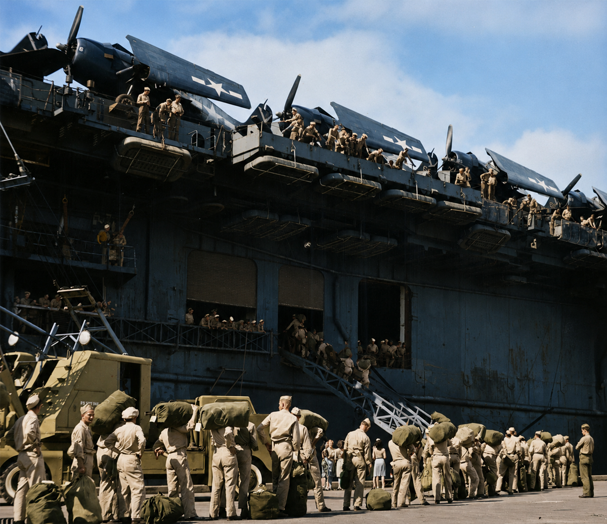Soldiers in uniform carry duffel bags and line up beside a large battleship, while more personnel and aircraft are visible on the ship’s deck under a blue sky.