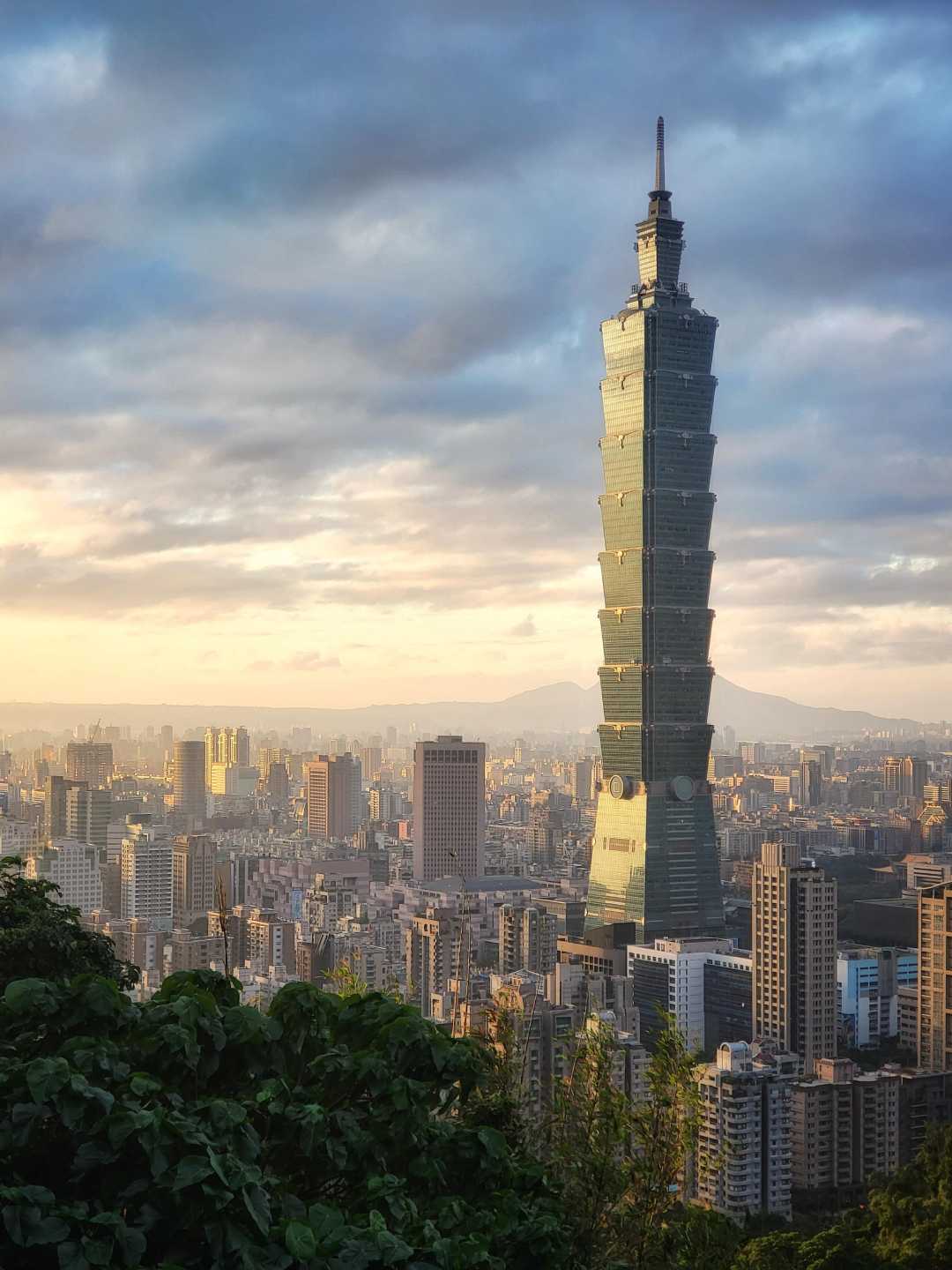 Skyscraper Taipei 101 towers over a cityscape of modern buildings at sunrise or sunset, with clouds in the sky and leafy greenery in the foreground.