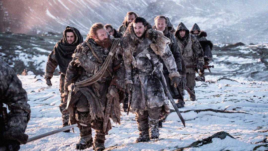 A group of people in rugged, fur-lined winter clothing walk across a snowy, rocky landscape with mountains in the background, looking serious and determined.