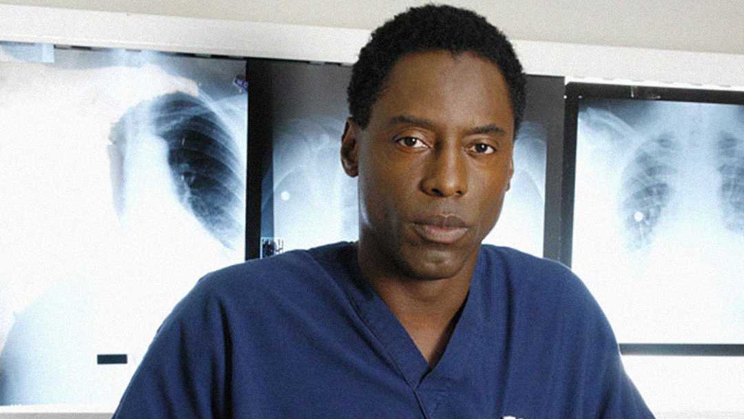 A man in blue medical scrubs stands in front of illuminated X-ray images, looking at the camera with a neutral expression.