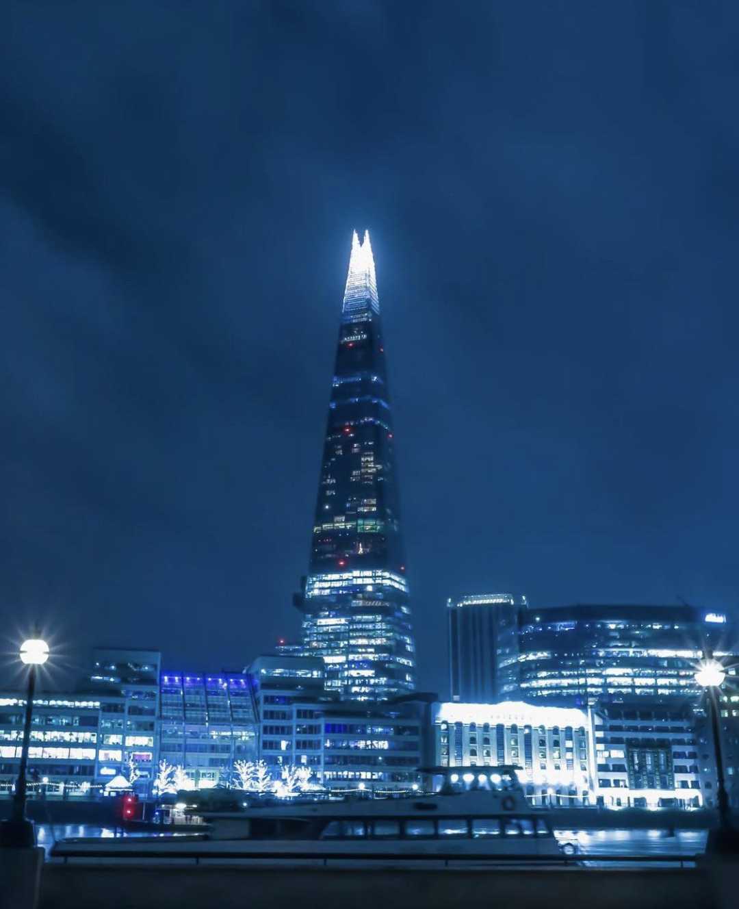 A nighttime view of the Shard skyscraper in London, illuminated with bright lights at the top, surrounded by other lit buildings under a dark, cloudy sky.