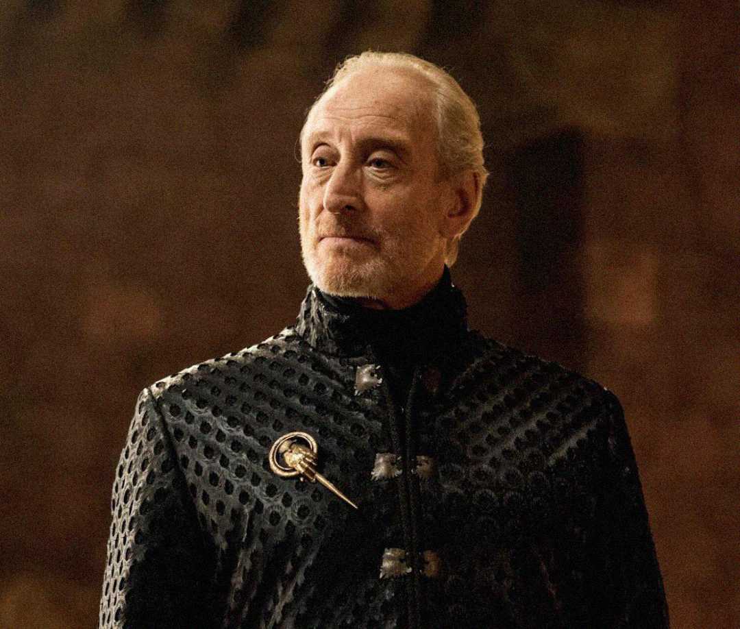 An older man with light hair and a beard wears a dark, textured jacket adorned with a gold brooch shaped like a hand, standing in a dimly lit room with a neutral expression.