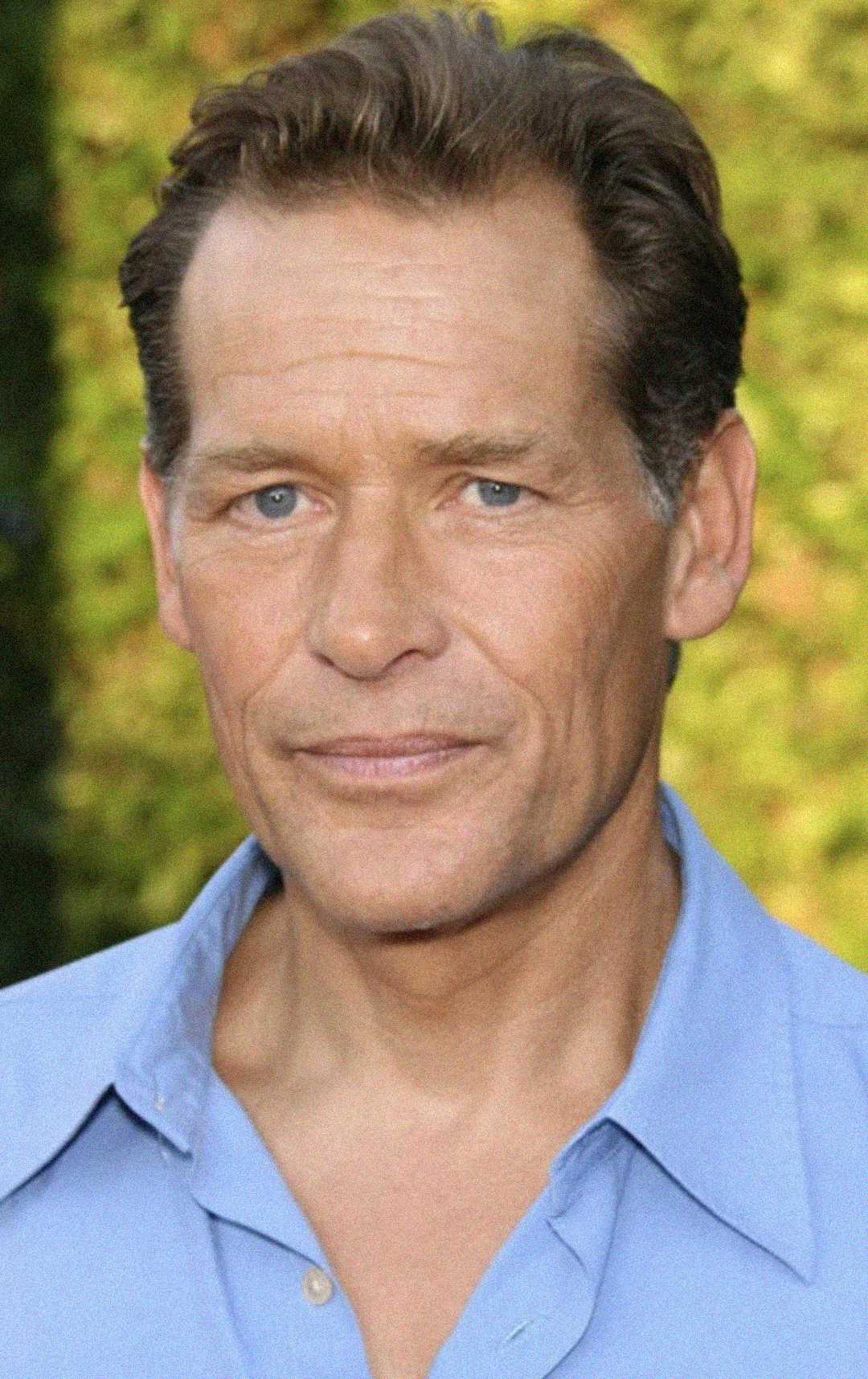 A middle-aged man with short brown hair and blue eyes is wearing a light blue collared shirt. He is standing outdoors in front of a blurred green background, looking directly at the camera with a neutral expression.