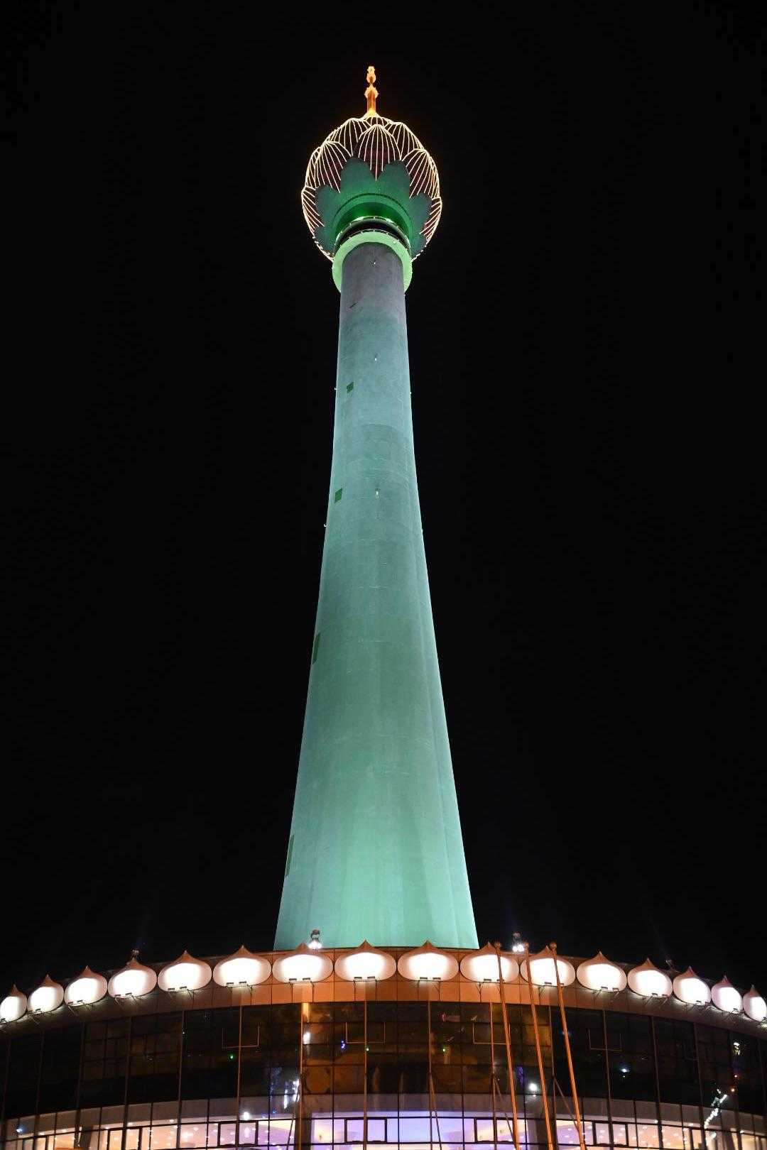 A tall communications tower illuminated in green and gold lights stands against a black night sky, with a distinctive dome structure at the top and a ring of lights along its base.