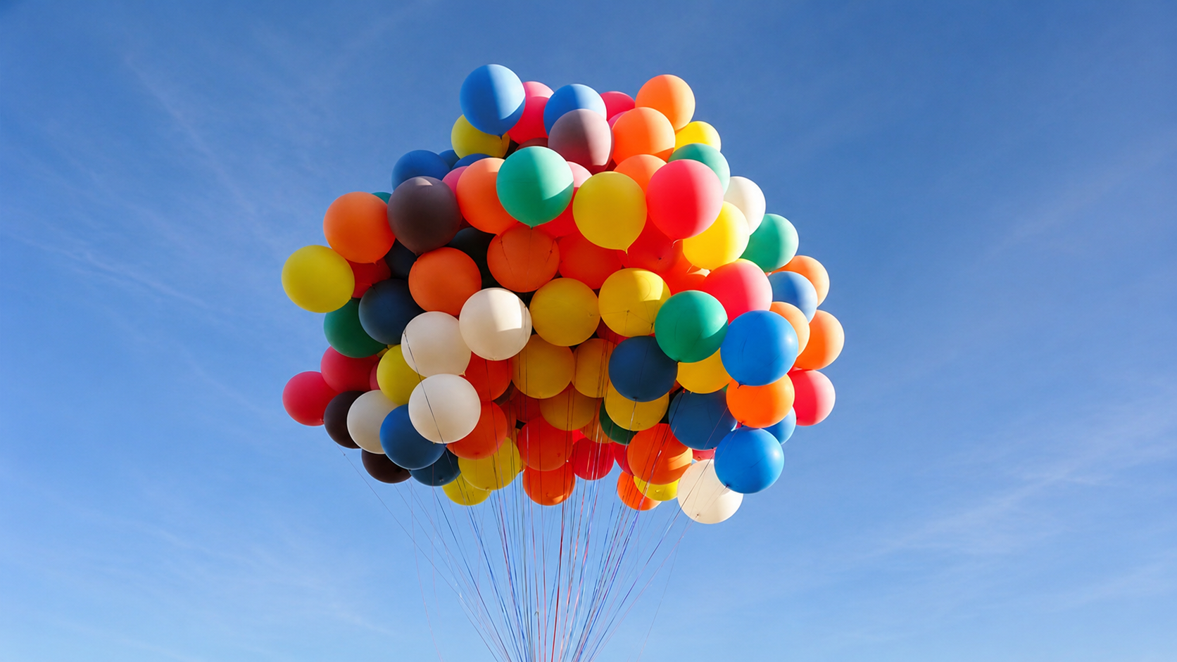A large bunch of colorful balloons, including red, yellow, blue, orange, white, and green, floating against a clear blue sky.