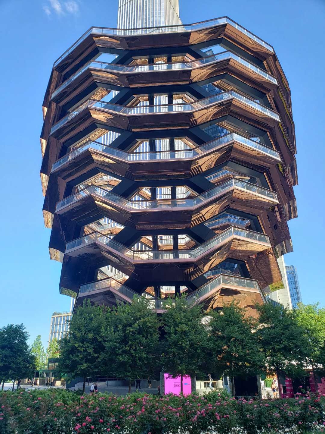 A large, honeycomb-like structure with multiple staircases, platforms, and terraces stands in a cityscape, surrounded by trees and greenery under a clear blue sky.
