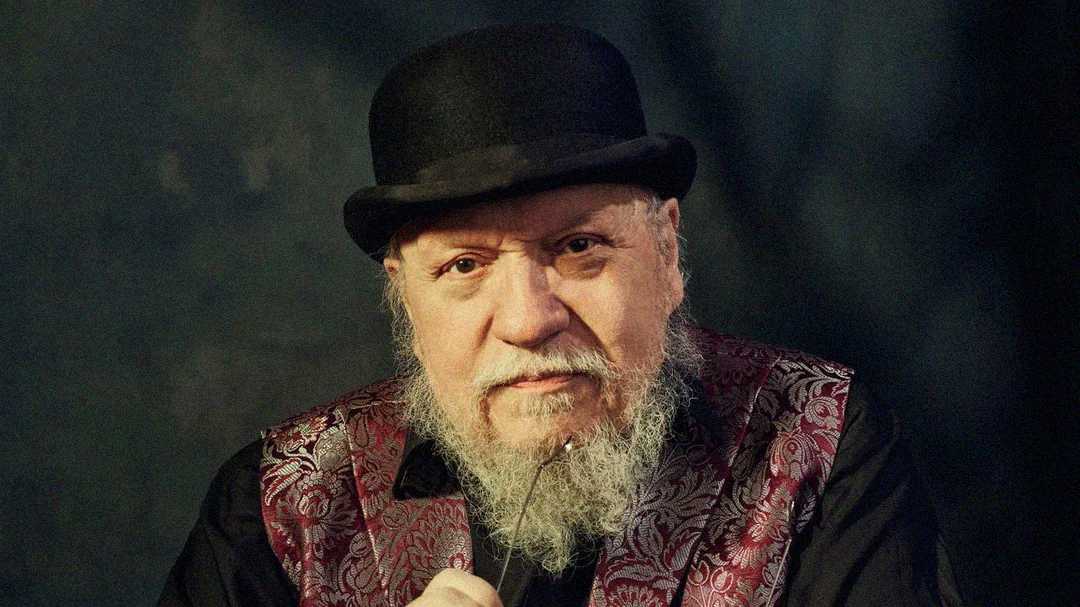 An older man with a gray beard wearing a black bowler hat and a dark shirt with a patterned maroon vest, gazing forward and holding a pair of eyeglasses. The background is dark and blurred.