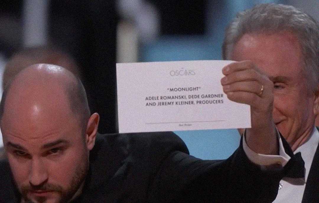A man holds up an Oscars envelope showing "Moonlight" as the winner, with names of producers Adele Romanski, Dede Gardner, and Jeremy Kleiner visible. Another man smiles in the background.