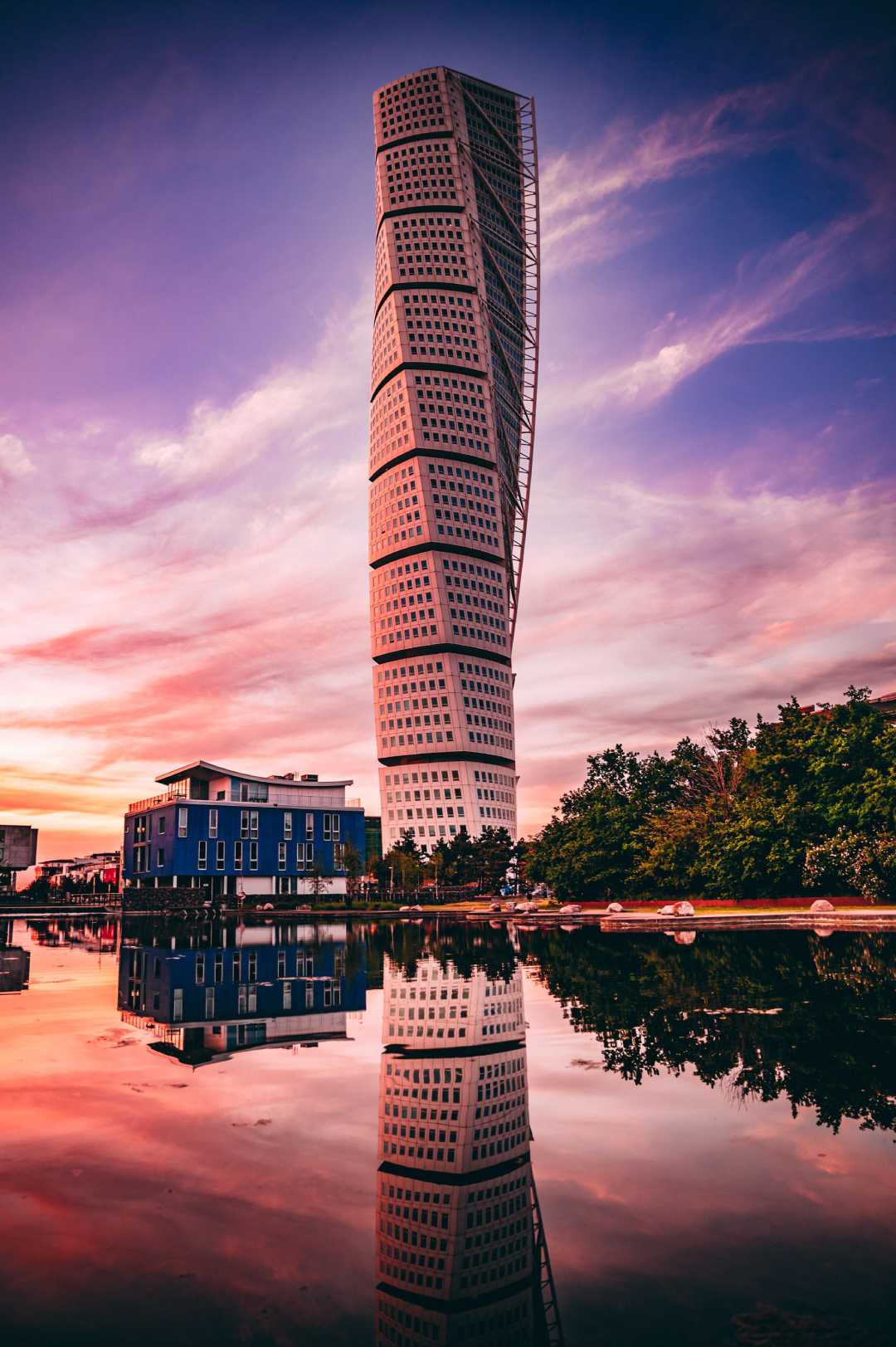 The Turning Torso skyscraper in Malmö, Sweden, stands tall against a vibrant sunset sky, its unique twisted design reflected in calm water below, with trees and buildings nearby.