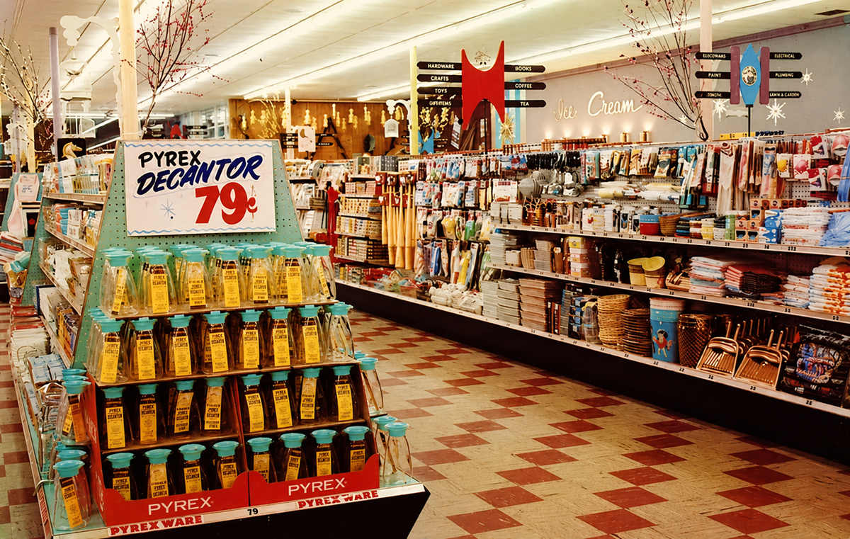 A vintage supermarket aisle displays Pyrex decanters for 79 cents on a central stand, surrounded by shelves of kitchenware, utensils, and home goods. The floor has a red and cream checkered pattern.