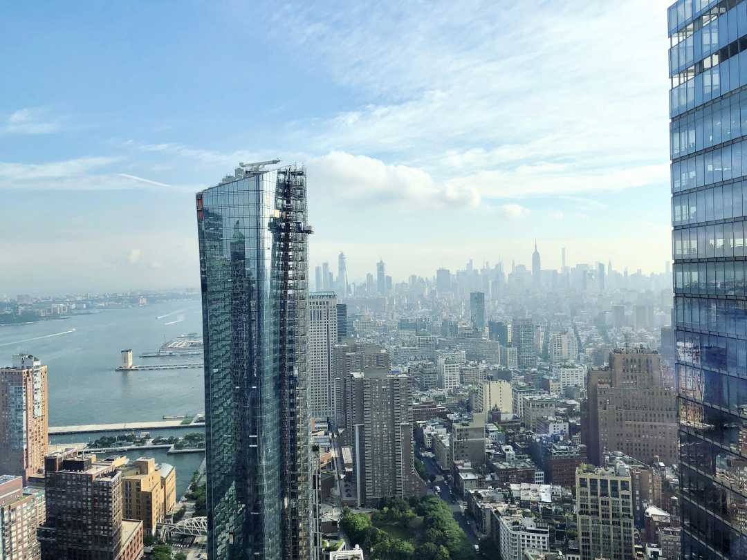 A hazy New York City skyline under a cloudy sky, with a modern glass skyscraper in the foreground reflecting the urban landscape and a river to the left.
