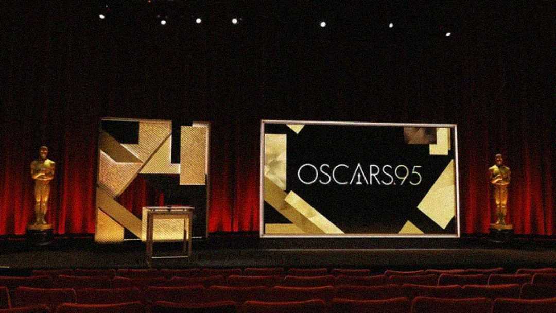 A stage with red curtains, two Oscar statues, and a large screen displaying "OSCARS 95" in gold and black geometric designs, viewed from the audience seating area.
