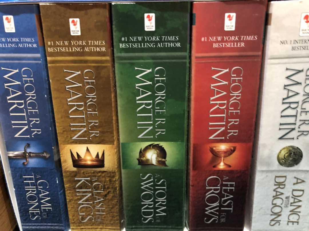 Five George R. R. Martin books from the A Song of Ice and Fire series on a shelf, including "A Game of Thrones," "A Clash of Kings," "A Storm of Swords," "A Feast for Crows," and "A Dance with Dragons.