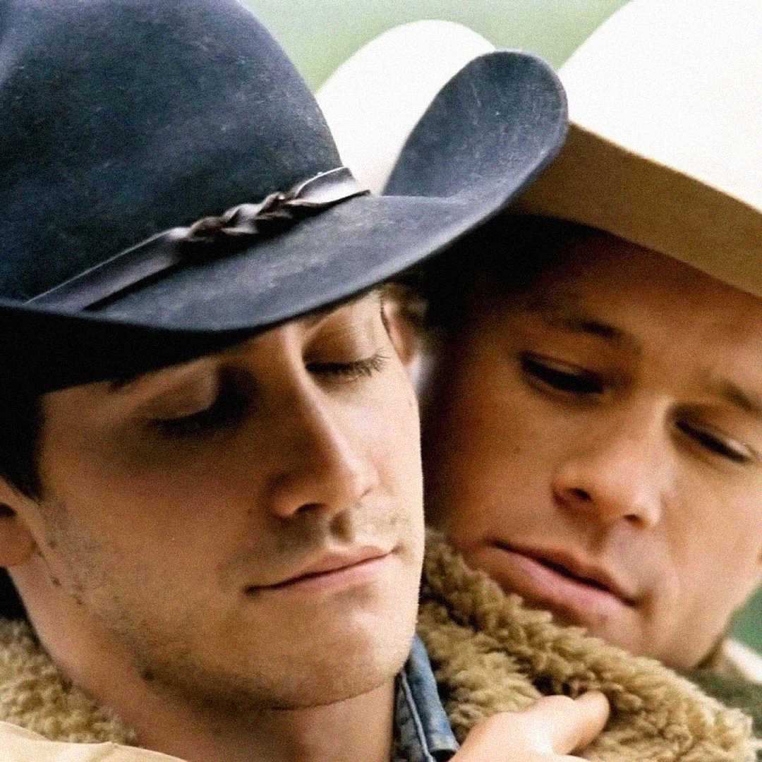 Two men wearing cowboy hats embrace closely, their faces touching gently. One man has his eyes closed, while the other looks down, creating an intimate and tender moment.