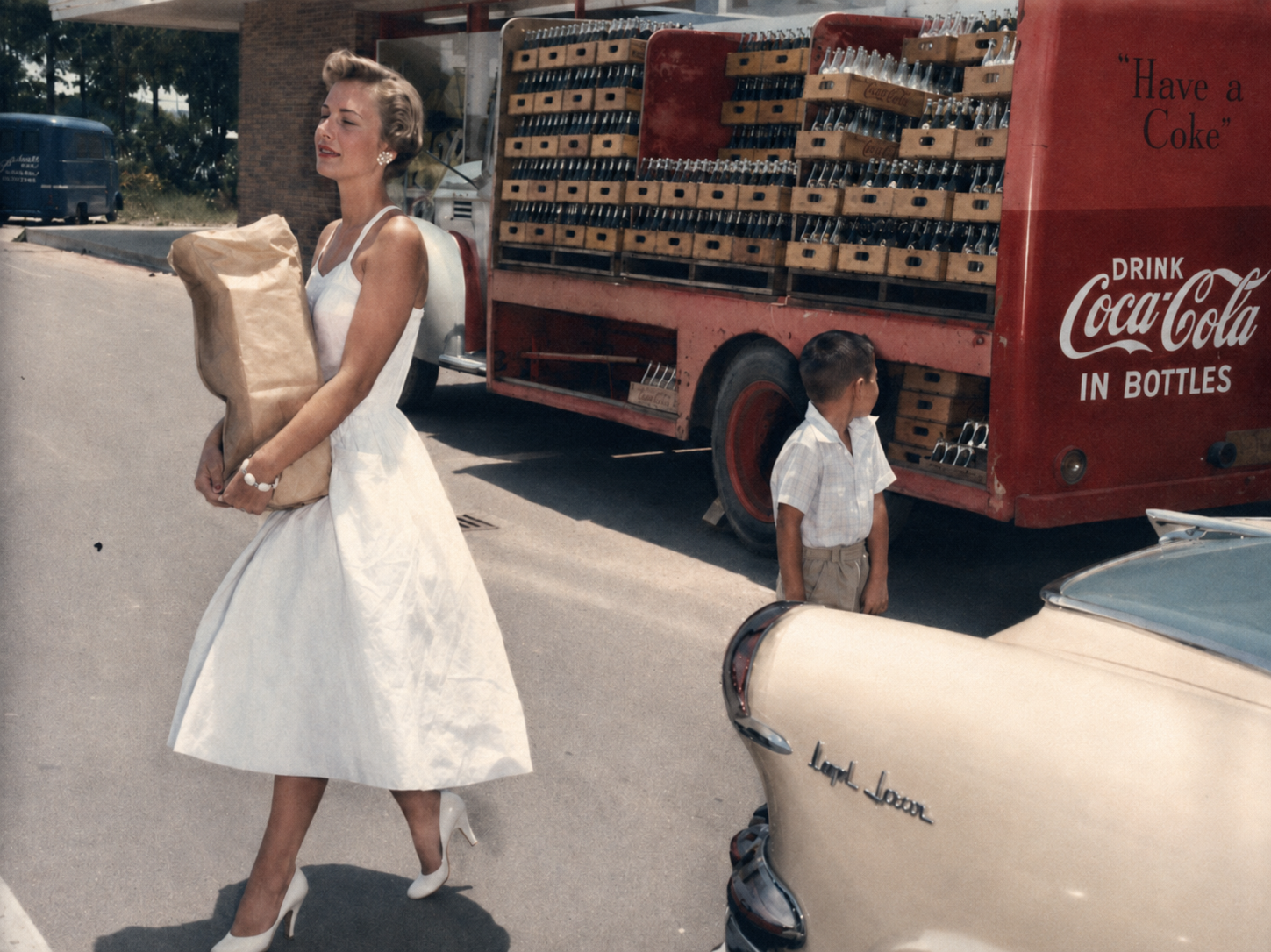 A woman in a white dress and heels carries a grocery bag past a young boy and a vintage Coca-Cola delivery truck loaded with crates. A cream-colored car with "Land Rover" on the back is parked nearby.