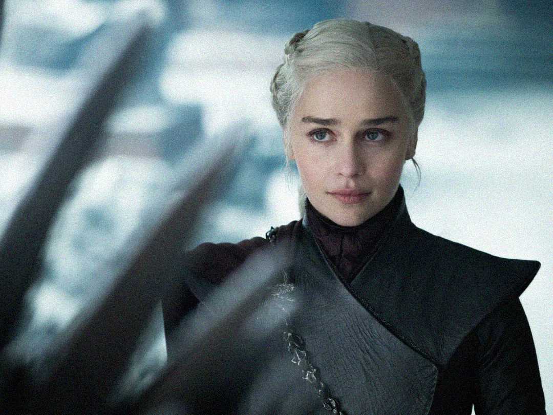 A woman with silver-blonde braided hair, dressed in dark medieval-style clothing, stands in front of blurred dragon-like spikes, gazing ahead with a serious expression. The background is soft and cool-toned.