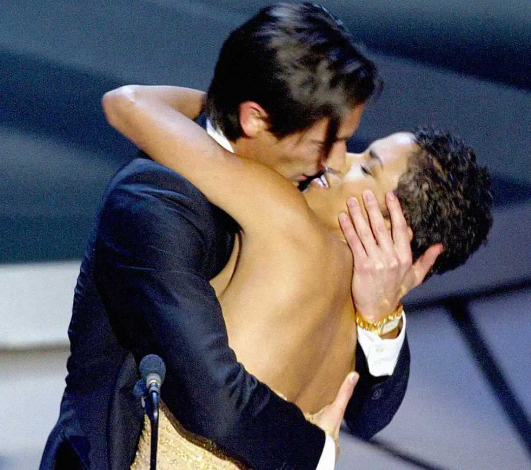 A man in a suit passionately hugs and kisses a woman in a strapless dress on stage, with a microphone visible in the foreground.