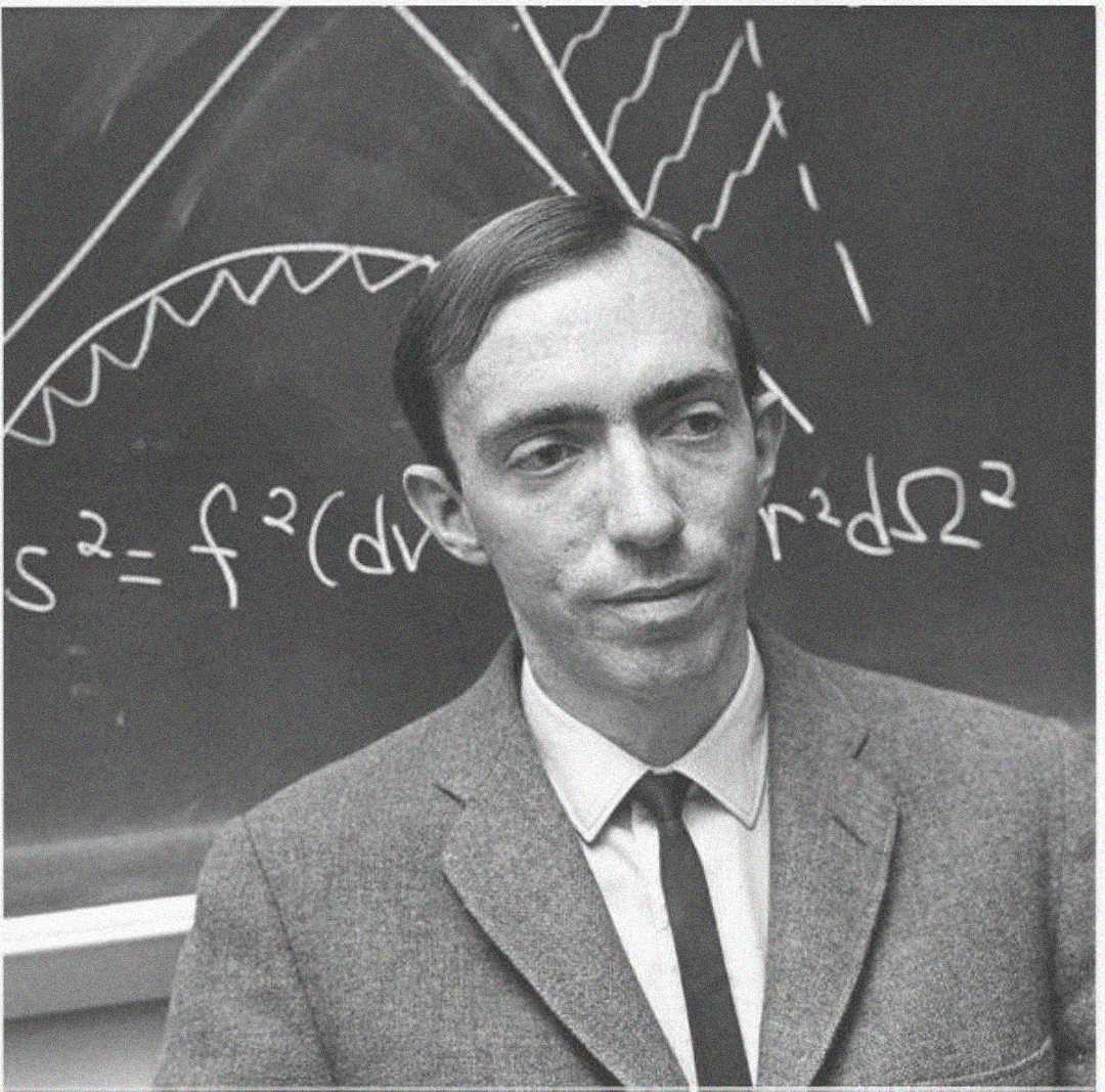 A man in a suit and tie stands in front of a chalkboard covered with mathematical equations and diagrams. He has short, dark hair and a neutral expression.