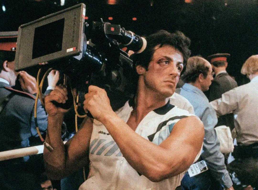 A man with a bruised face and muscular arms holds a large film camera on his shoulder in a crowded indoor setting, wearing a sleeveless shirt with sweat marks. People are gathered in the background.