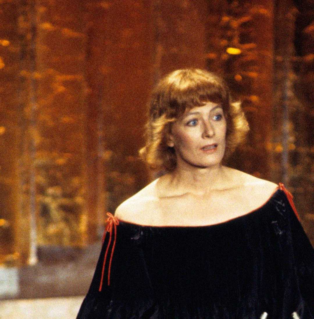 A woman with short, curly reddish hair stands in front of a blurred golden background, wearing an off-the-shoulder black dress with red ties on the sleeves. She appears to be speaking or reacting to something.