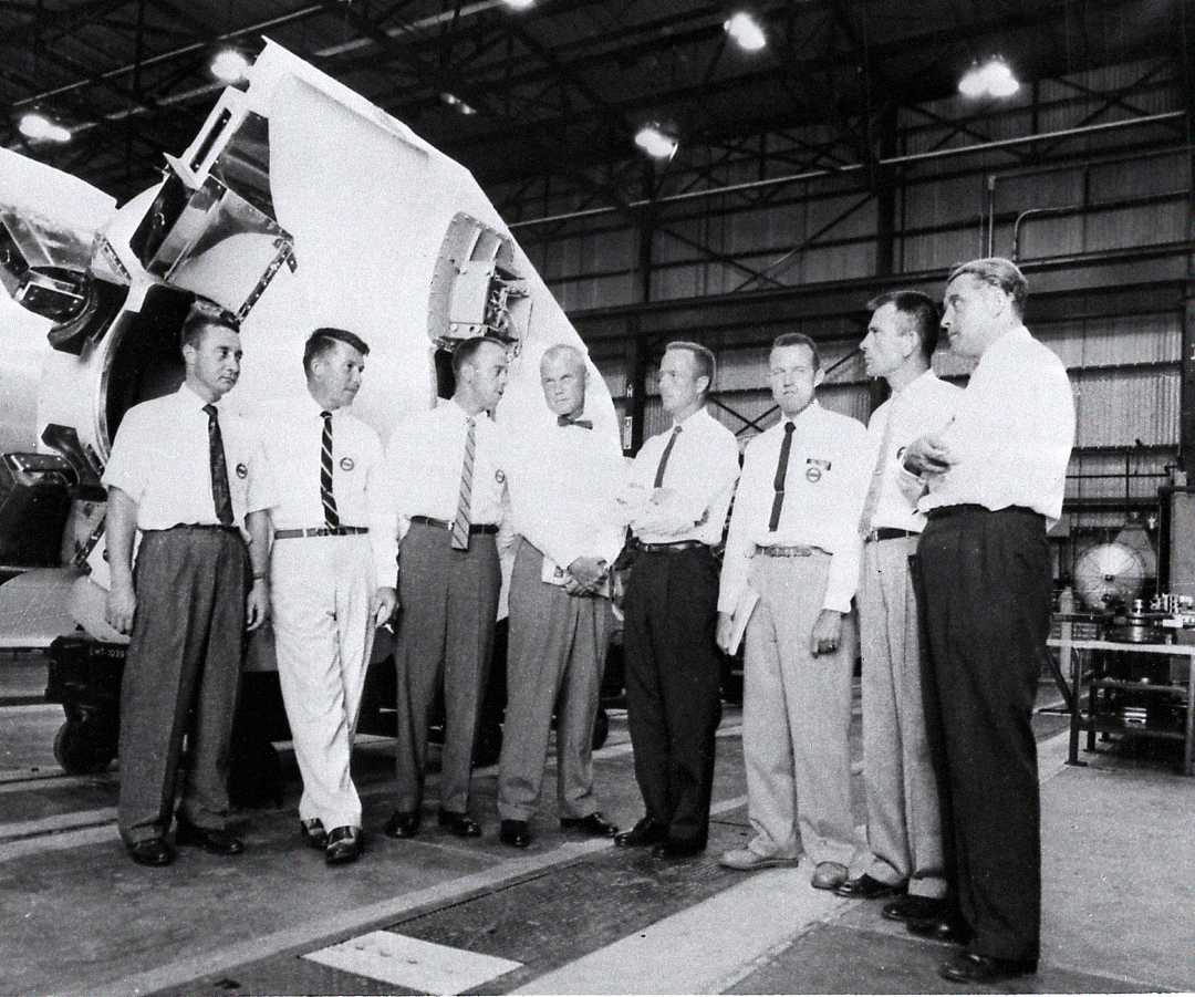 Seven men in dress shirts and slacks stand together in a hangar, conversing in front of a large spacecraft part. The background is industrial, with visible beams and equipment.