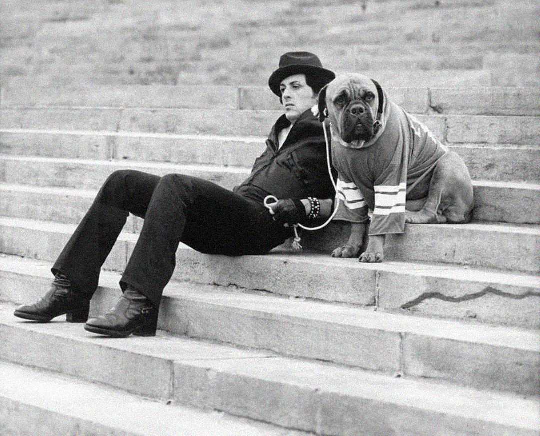 A person in dark clothing and a hat sits on stone steps, holding a leash attached to a large dog wearing a sweater. Both appear relaxed, looking in the same direction. The image is black and white.