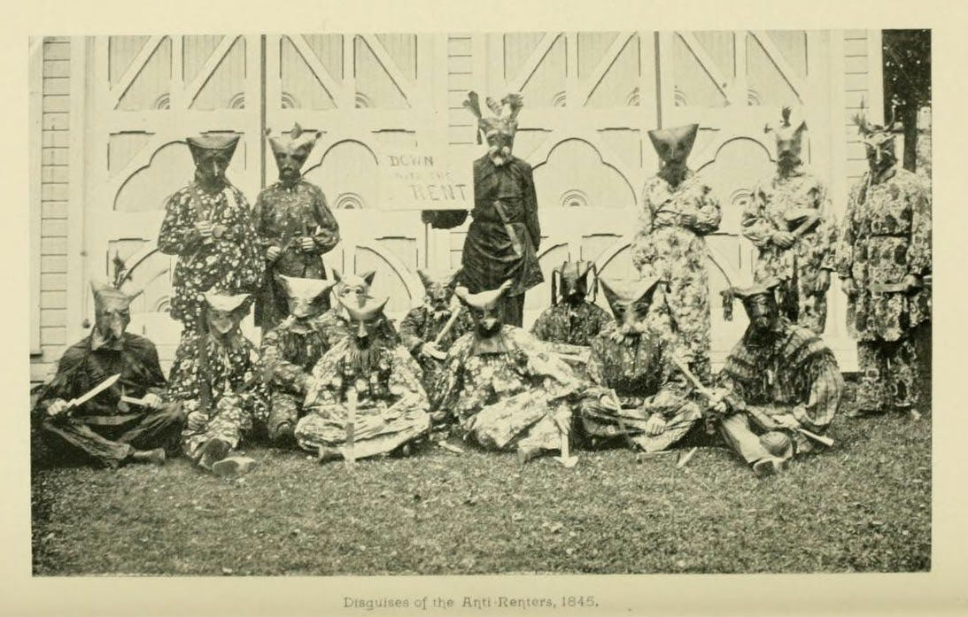 Historical black-and-white photo of thirteen people in patterned costumes and horned masks, posing in front of a building. A sign behind them reads “DOWN WITH RENT.” Caption: “Disguises of the Anti-Renters, 1845.”