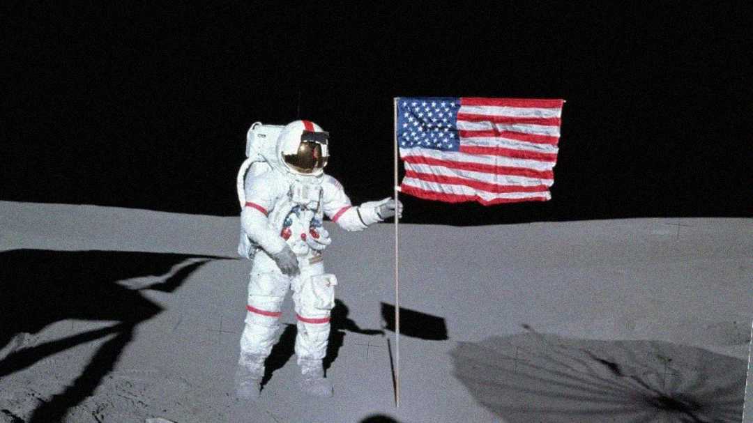 An astronaut in a white spacesuit stands on the moon's surface holding an American flag. The sky is black, and lunar soil stretches in the background.