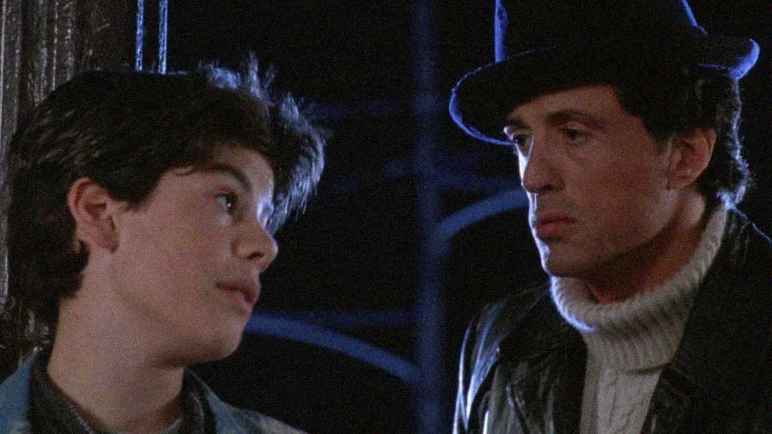 A boy with dark hair looks up at a man in a dark hat and turtleneck sweater. The man gazes at the boy seriously. It is nighttime and both appear to be outdoors.