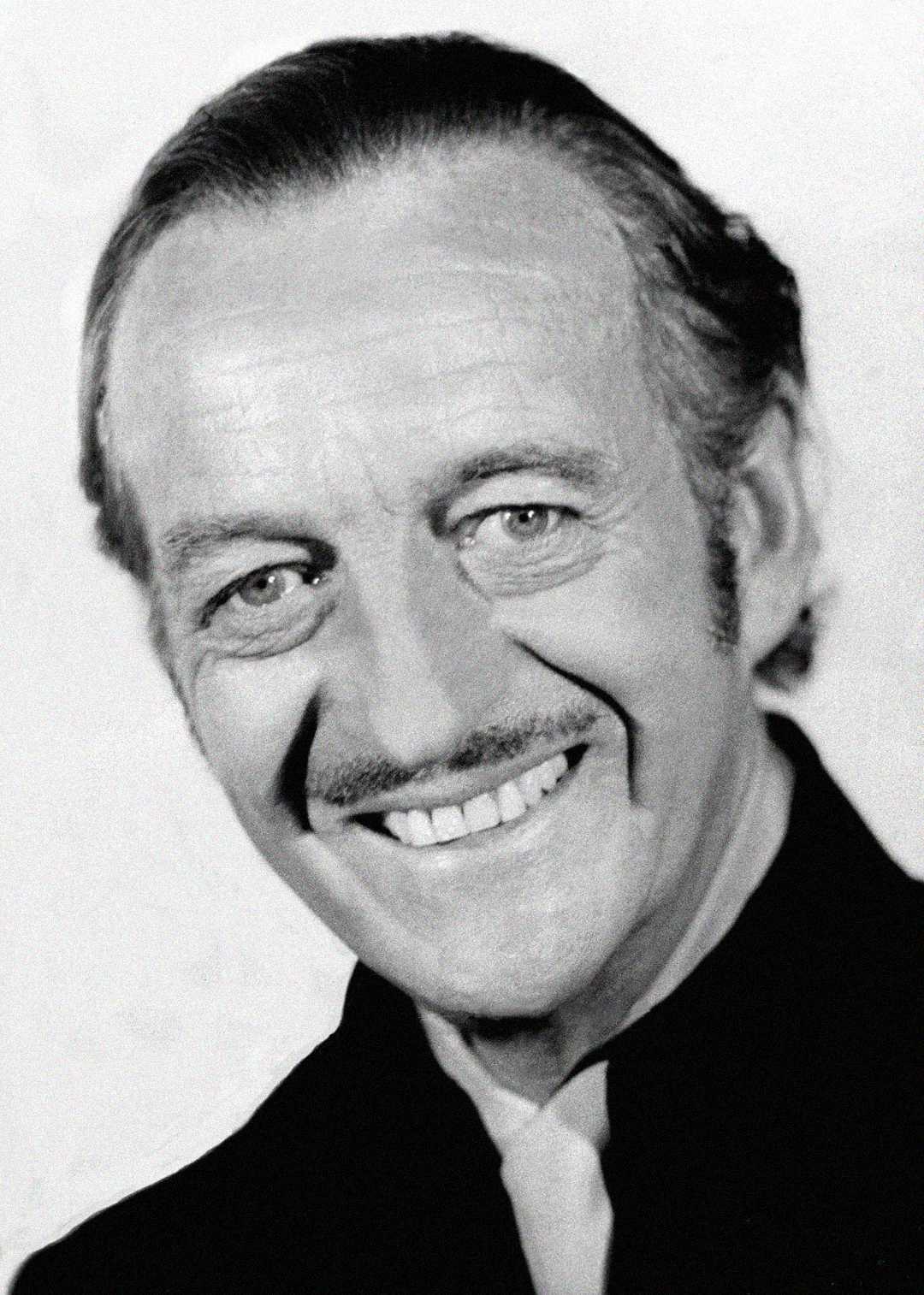 A black and white portrait of an older man with slicked-back hair, a thin mustache, and a broad smile, wearing a dark jacket and light shirt, looking directly at the camera.
