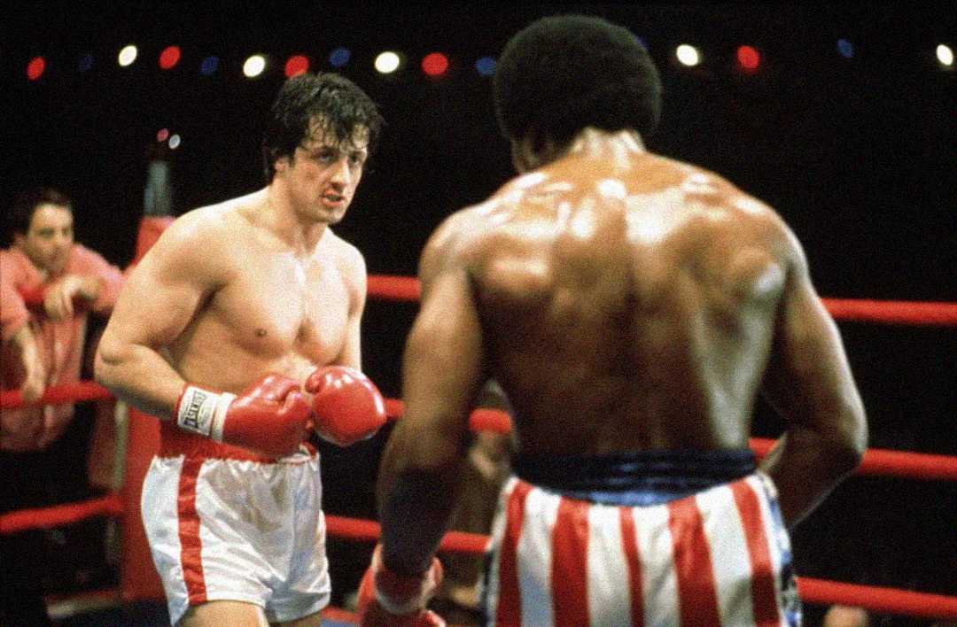 Two male boxers face each other in a boxing ring; one with red gloves and white trunks with a red stripe, and the other with his back to the camera, wearing red, white, and blue striped trunks.
