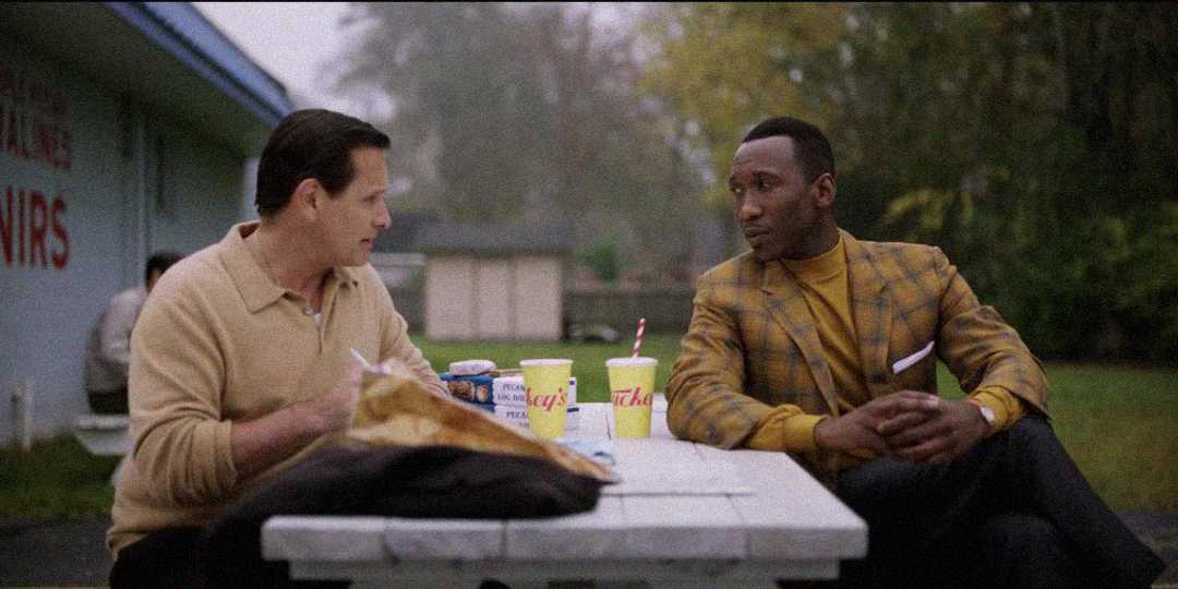 Two men sit across from each other at a picnic table outside, talking. Fast food items and drinks are on the table. One man wears a beige sweater; the other wears a yellow plaid jacket. Trees and a building are in the background.