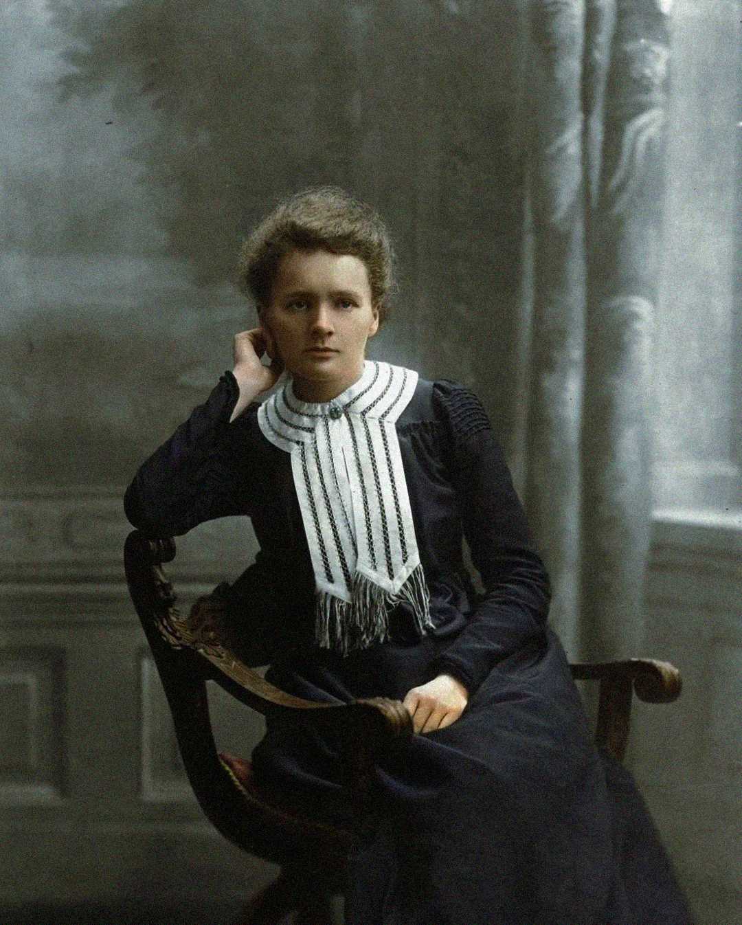 A young woman with short, curly hair sits on a wooden chair, wearing a dark dress with a distinctive white, pleated collar. She rests her head on one hand and looks directly at the camera, with a neutral expression.