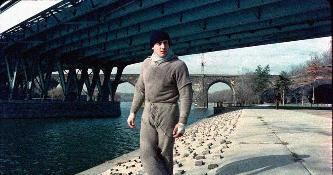 A man in gray workout clothes and a black beanie walks along a riverbank under a large bridge, with stone arches and trees in the background.