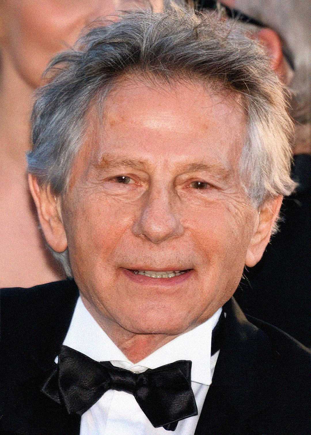 An older man with gray hair wearing a black tuxedo and bow tie, smiling slightly at the camera. The background is blurred with indistinct people.