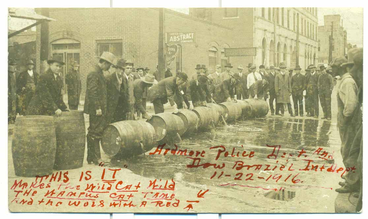 A sepia-toned photo shows a group of men in hats and coats standing around barrels lined up on a wet street. Red handwritten text is scrawled across the image with buildings visible in the background.