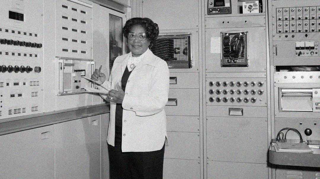 A woman in a lab coat stands in front of vintage computer equipment, holding a clipboard and pen, smiling as she works in a control room filled with buttons, panels, and switches.
