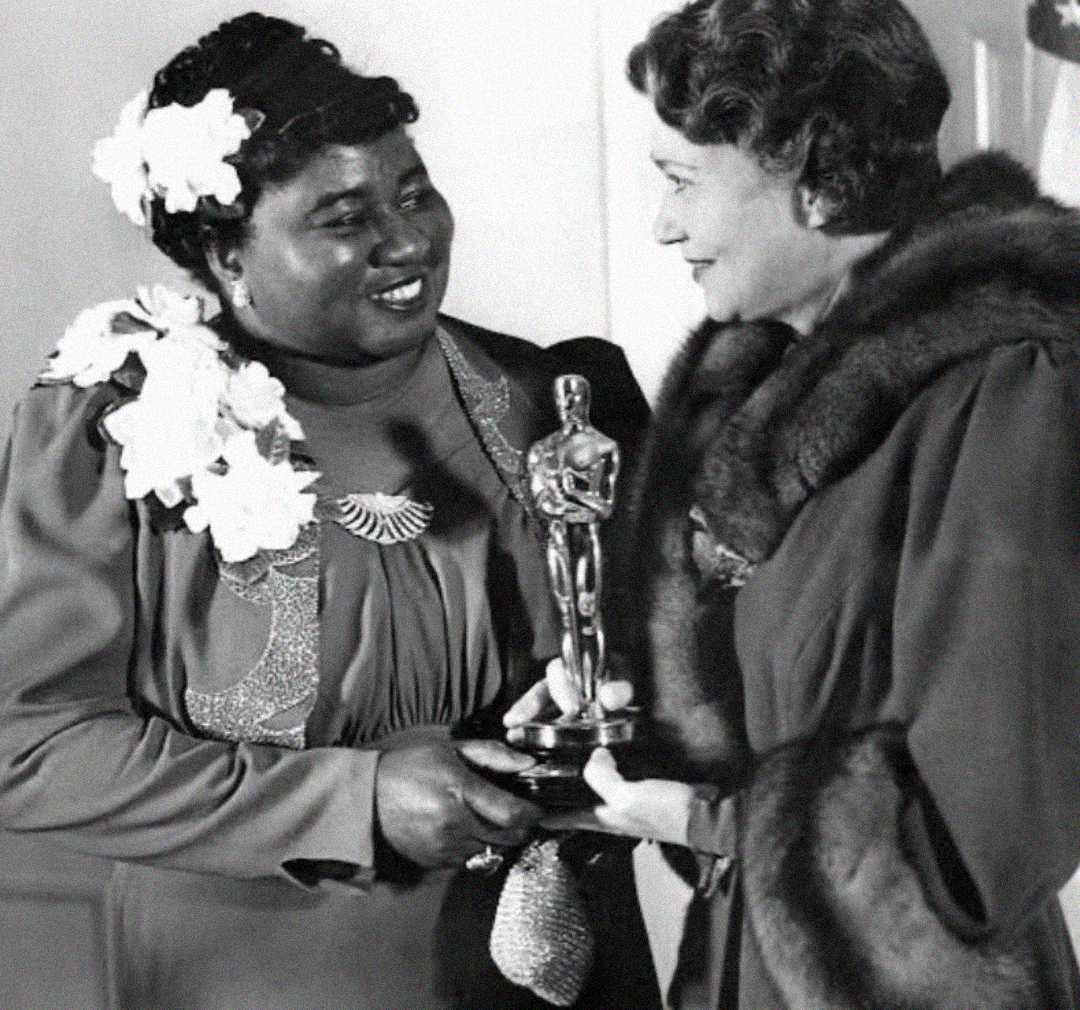 Two women stand together; one, in a floral-adorned dress, smiles while holding an Oscar statuette, as the other, wearing a fur-collared coat, presents it to her. The moment appears celebratory and formal.