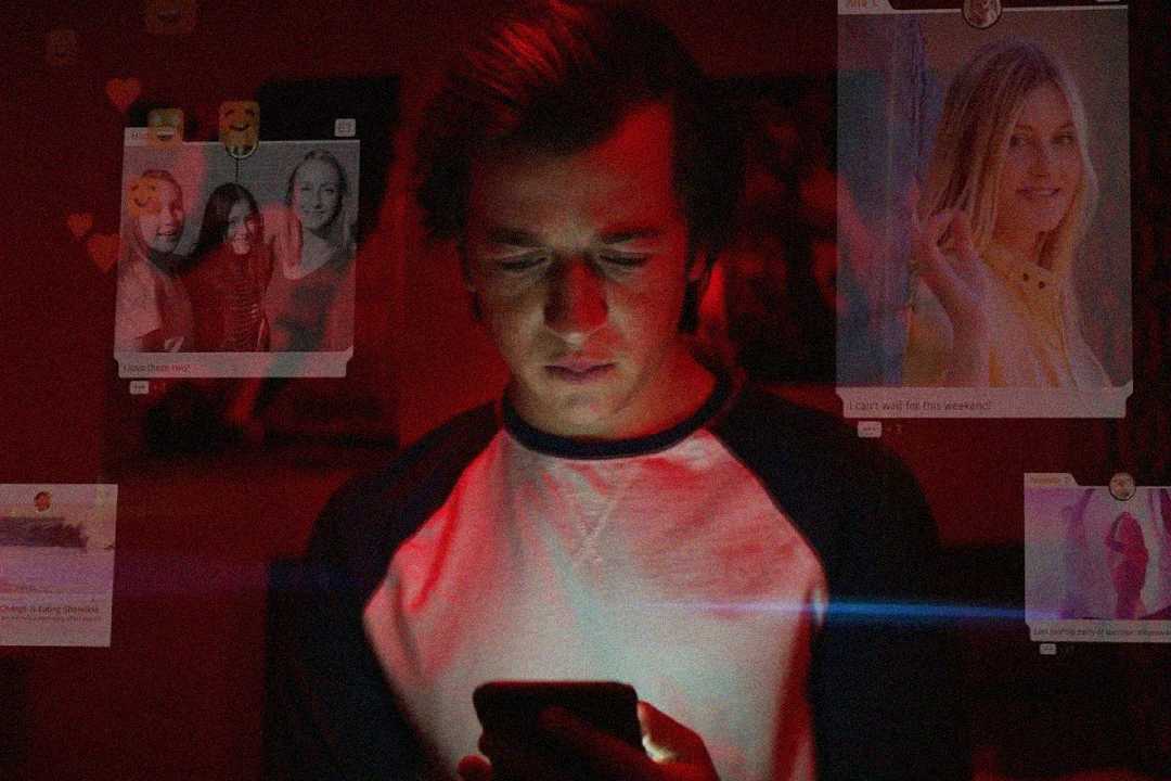 A young person stands in a dimly lit room, looking down at their phone. Social media posts and profile images are digitally overlaid around them, illuminating their face with a reddish glow.