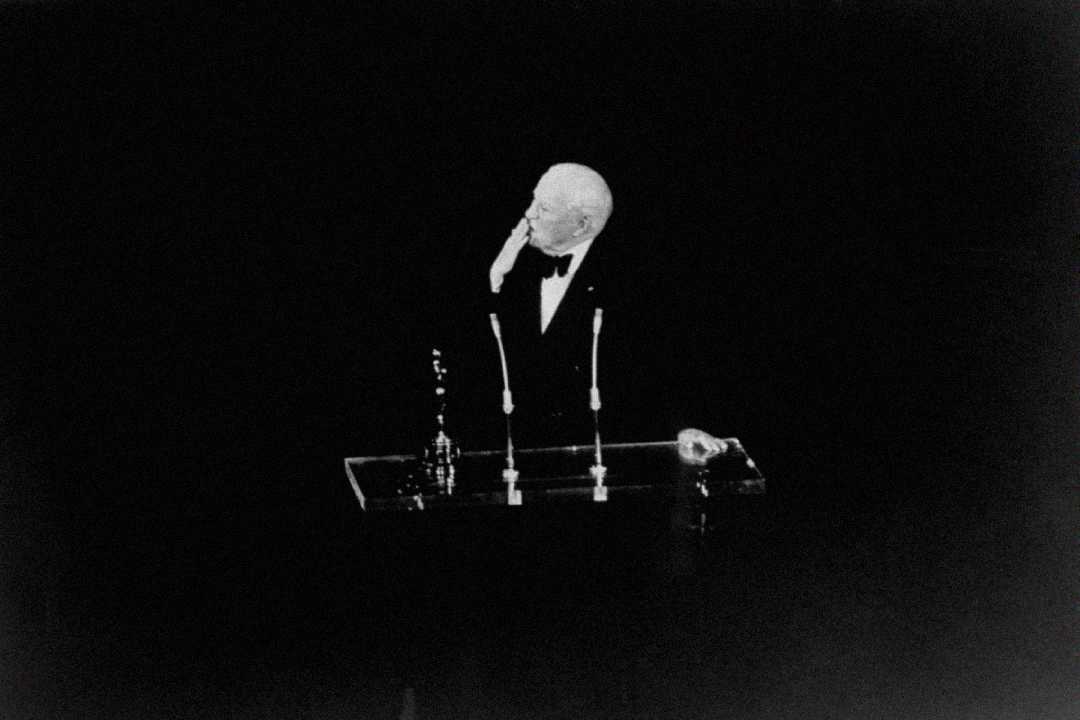 A man in a tuxedo stands alone at a podium with two microphones on a dark stage, holding his hand to his face in a thoughtful gesture.